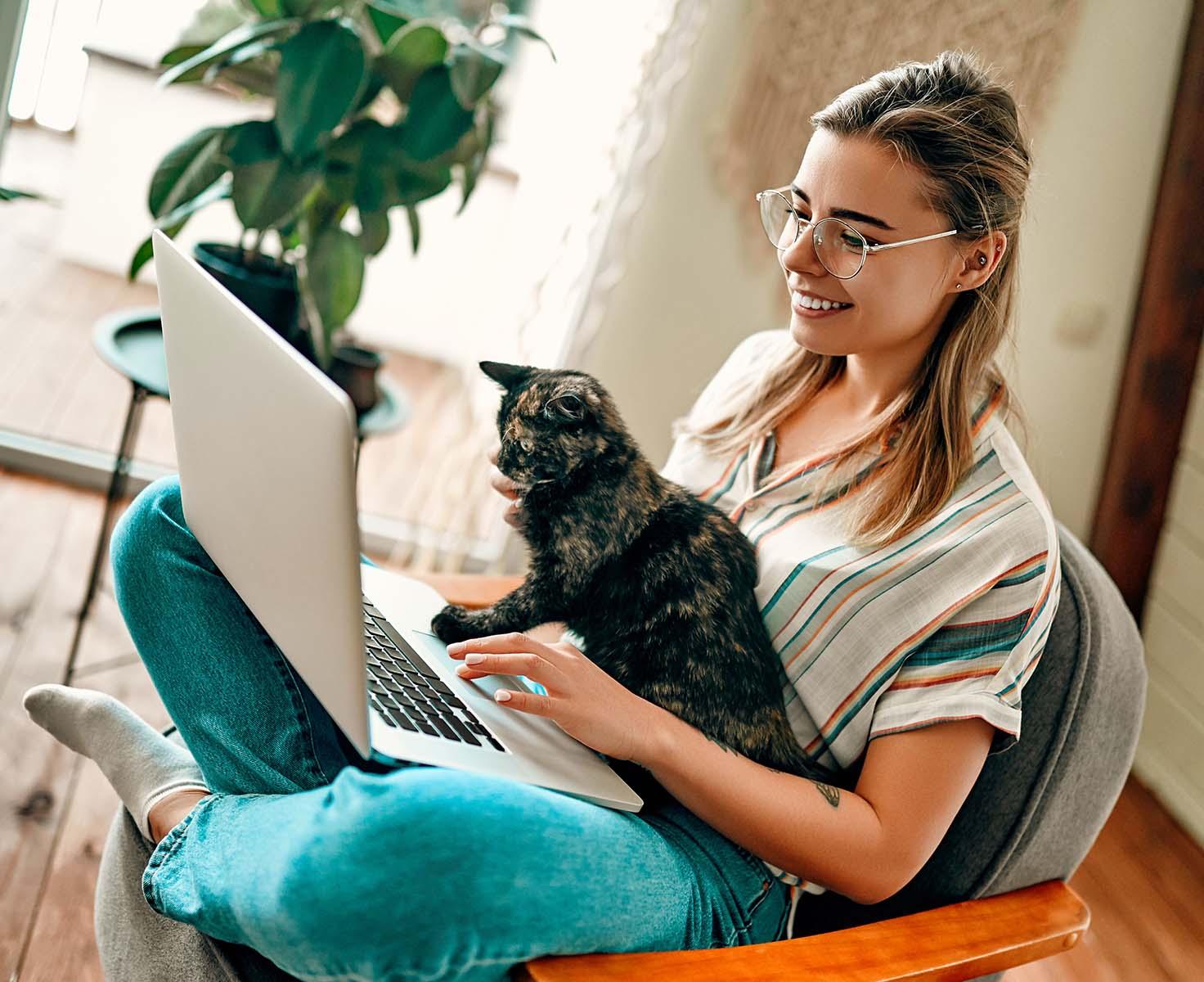 一位戴眼镜的女士和她的猫在笔记本电脑上工作时微笑着